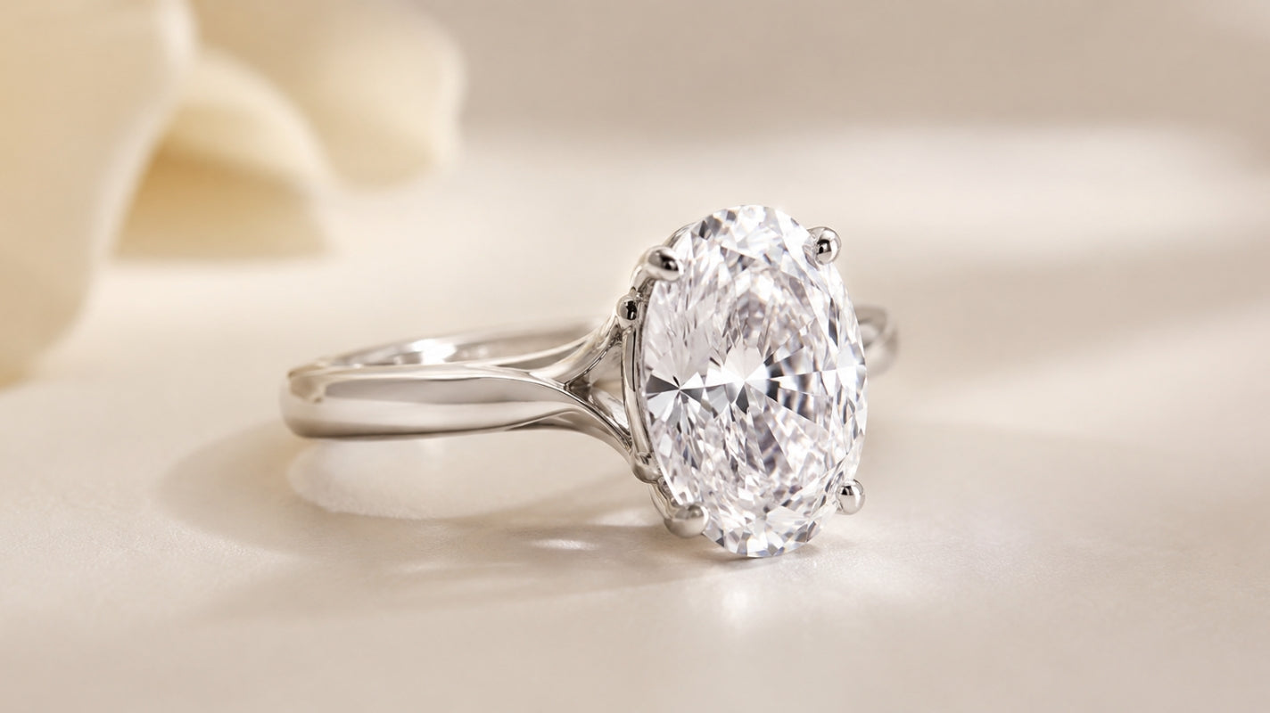 Elongated oval diamond solitaire in a polished white-metal custom setting, photographed at a three-quarter angle on a soft champagne background.