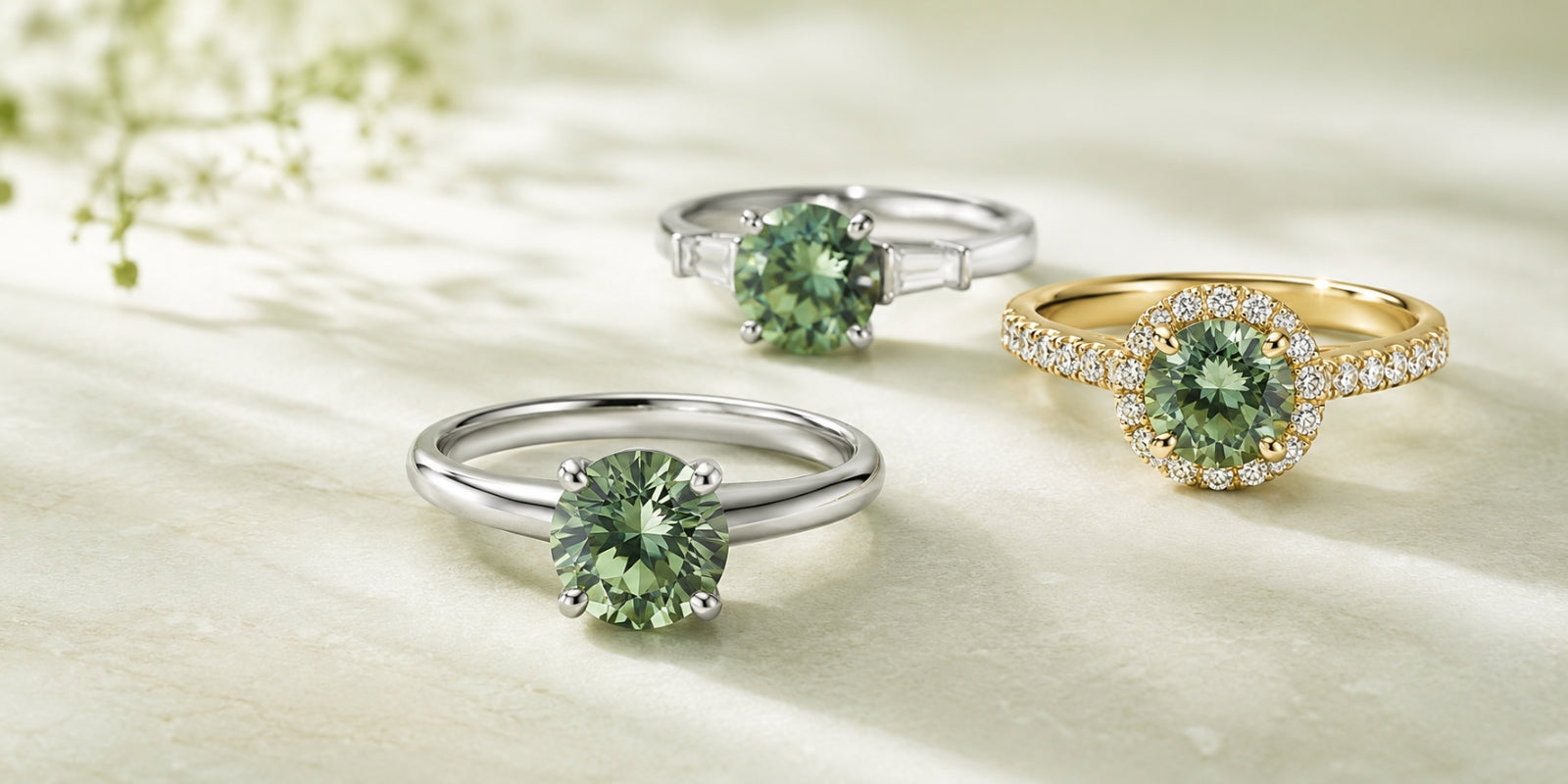 Three lab-grown green diamond rings displayed on a softly lit stone surface: a white-metal solitaire, a white-metal ring with baguette side stones, and a yellow-gold halo ring.
