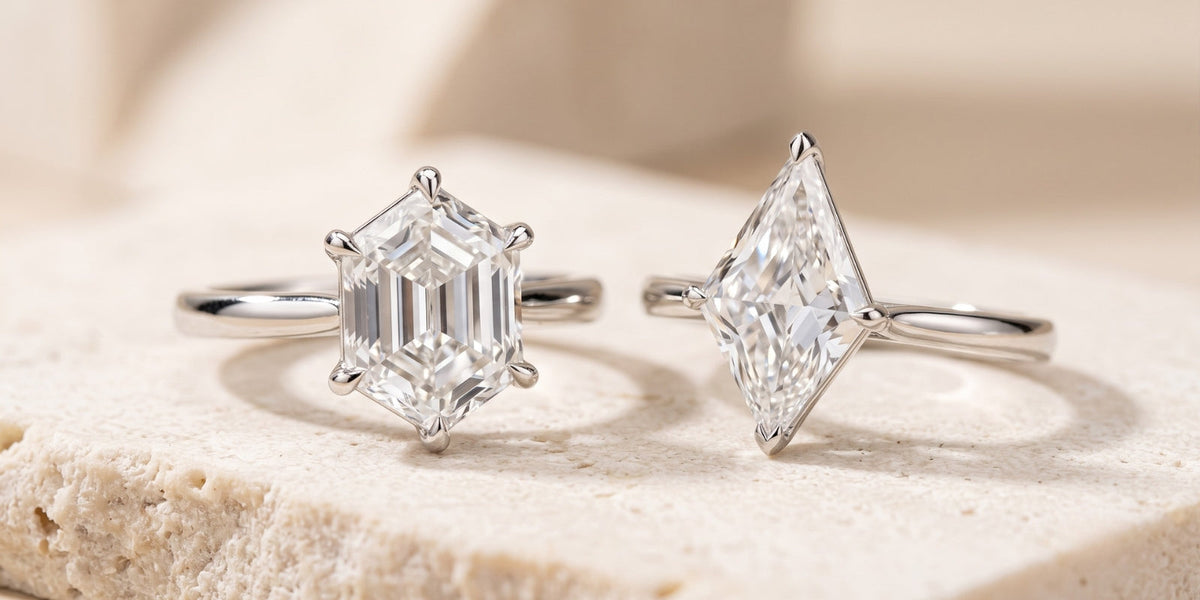 Luxury macro photograph of two white-metal engagement rings resting on warm ivory stone: one with an elongated hexagon step-cut diamond and one with a pointed kite-shaped brilliant diamond.
