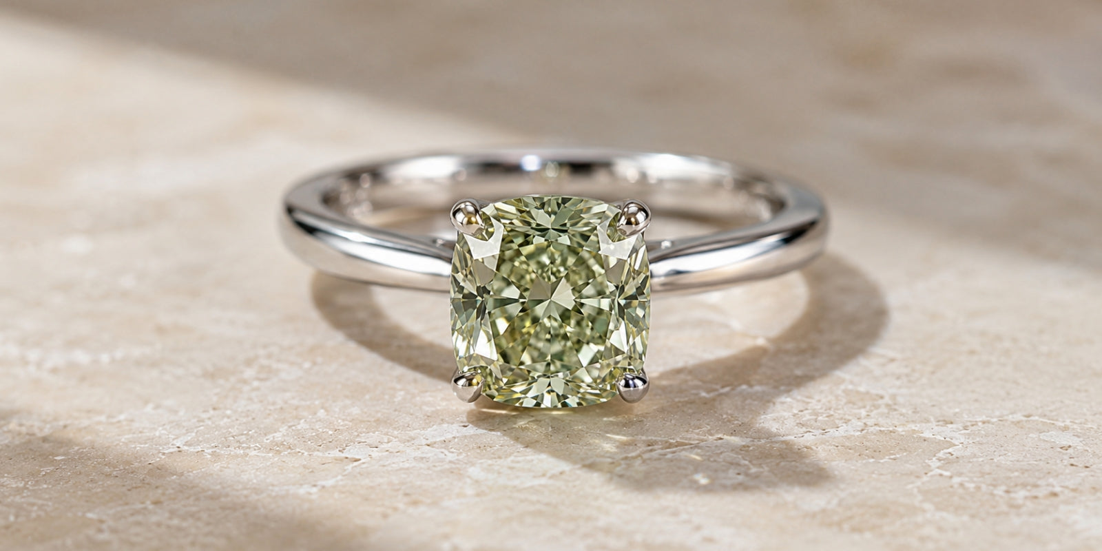 Close-up of a cushion-cut light green diamond set in a four-prong solitaire ring on a neutral stone surface.