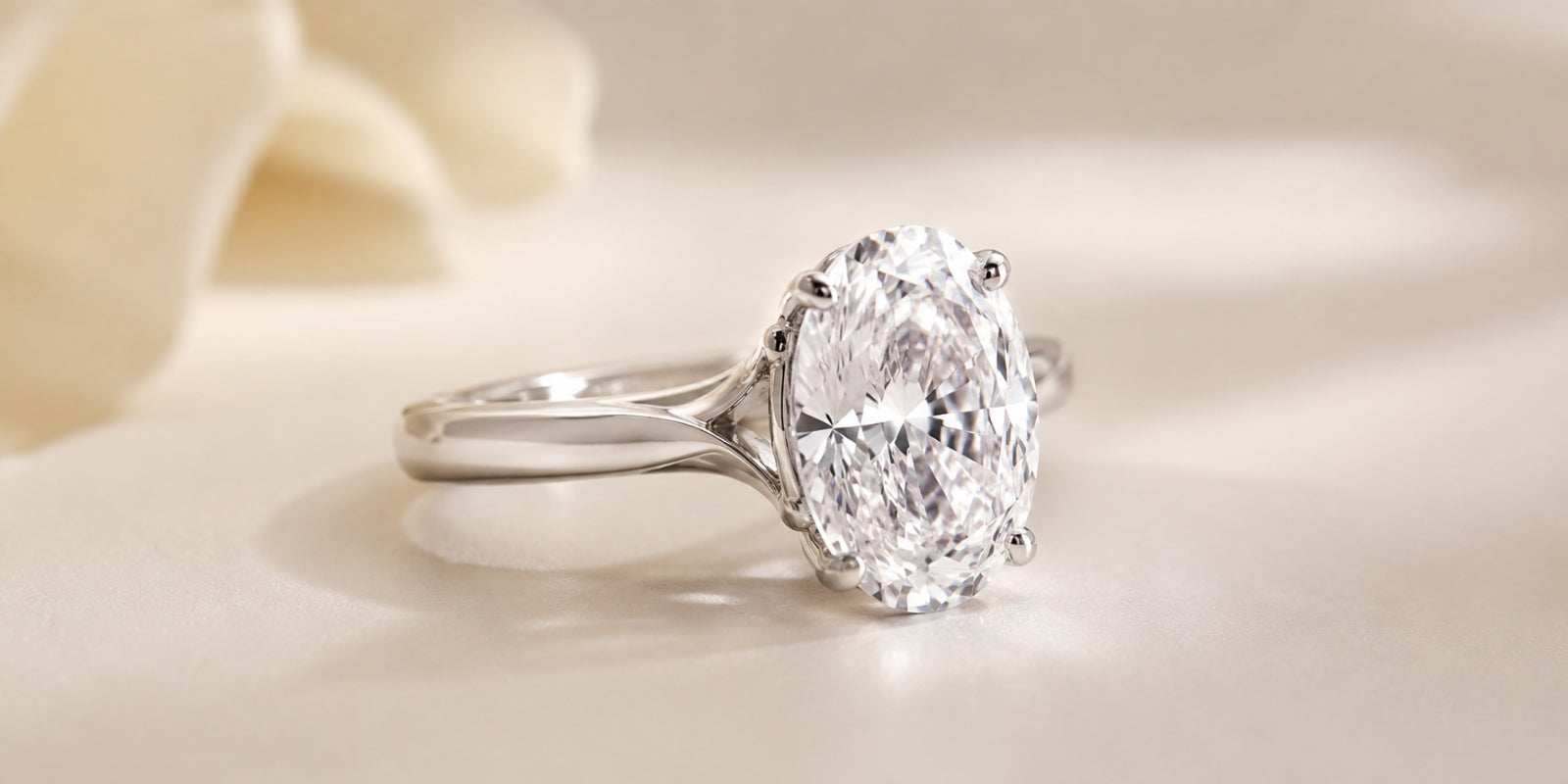 Elongated oval diamond solitaire in a polished white-metal custom setting, photographed at a three-quarter angle on a soft champagne background.